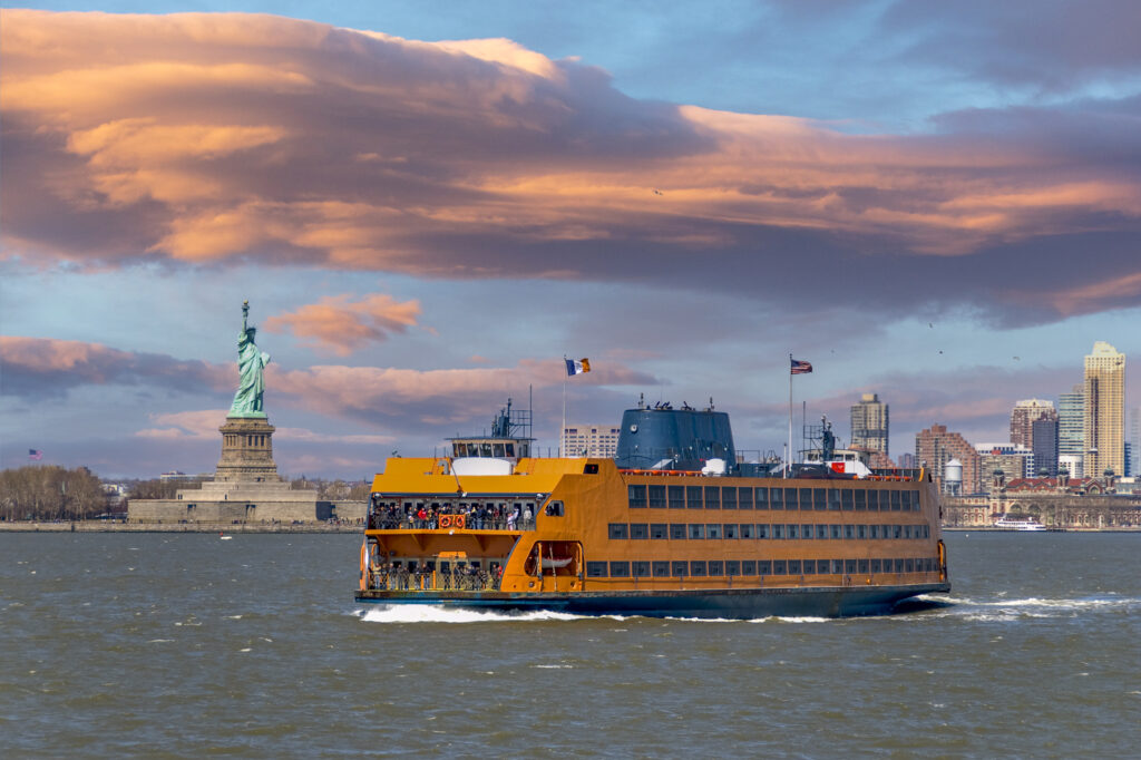 Staten Island ferry coming from Manhattan and passing by the Statue of Liberty. The Statue of Liberty and the skyscrapers of lower manhattan can be seen in the background.